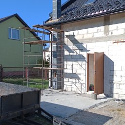 Robert Jórkowski - Budowa domu z białych bloczków, widoczne rusztowanie przy kominie, dach z czarnej dachówki. Przyczepka na trawie. Dom w trakcie budowy w słoneczny dzień.