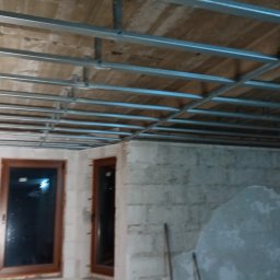 Robert Jórkowski - Montaż metalowej konstrukcji sufitu podwieszanego w surowym wnętrzu z oknami i ścianami z betonu komórkowego. Widoczne profile i elementy mocujące.