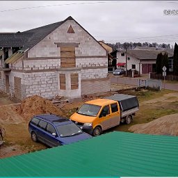 Robert Jórkowski - Budowa domu z białego pustaka, częściowo zadaszonego. Na posesji zaparkowane dwa samochody, widoczna betoniarka i hałdy piasku. Ujęcie z góry, z dachu sąsiedniego budynku.