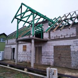 Robert Jórkowski - Budowa domu z bloczków: surowy stan z zieloną konstrukcją dachu. Widoczne bloczki, drewniane elementy i przygotowane materiały budowlane. Ujęcie z zewnątrz, w dzień.