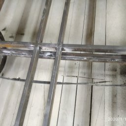 Robert Jórkowski - Spawana, metalowa konstrukcja ramy na drewnianej podłodze. Widoczne spawy i elementy zawieszenia. Ujęcie z dołu, podkreślające solidność wykonania.