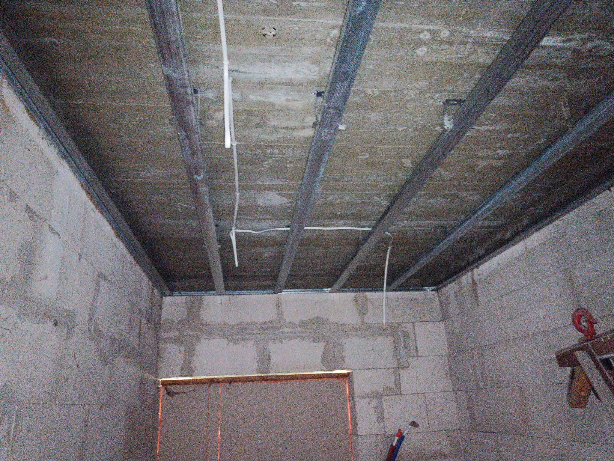 Montaż metalowej konstrukcji sufitu podwieszanego w surowym wnętrzu. Widoczne profile, przewody elektryczne i ściany z betonu komórkowego.