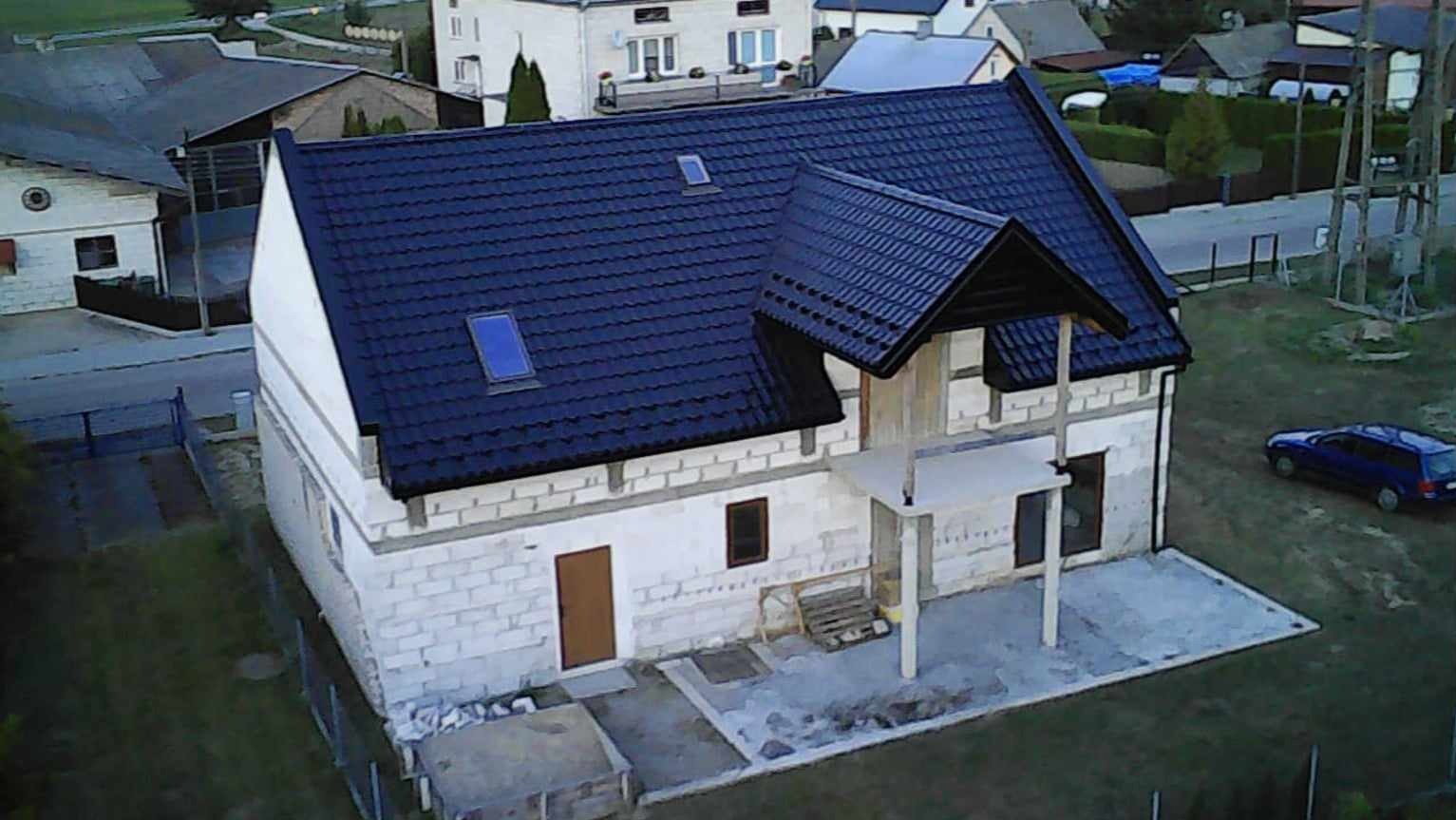 Niedokończony dom z ciemnym dachem i oknami dachowymi, widok z góry. Białe ściany z pustaków, widoczny fragment podwórka i samochód w tle.