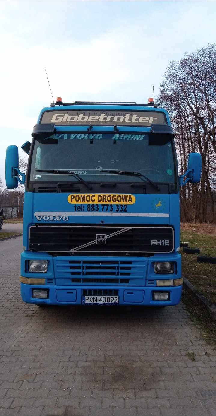 Błękitny samochód ciężarowy Volvo FH12 z napisem 'Pomoc Drogowa' na żółtym tle, zaparkowany na brukowanej nawierzchni. Widoczne drzewa w tle.