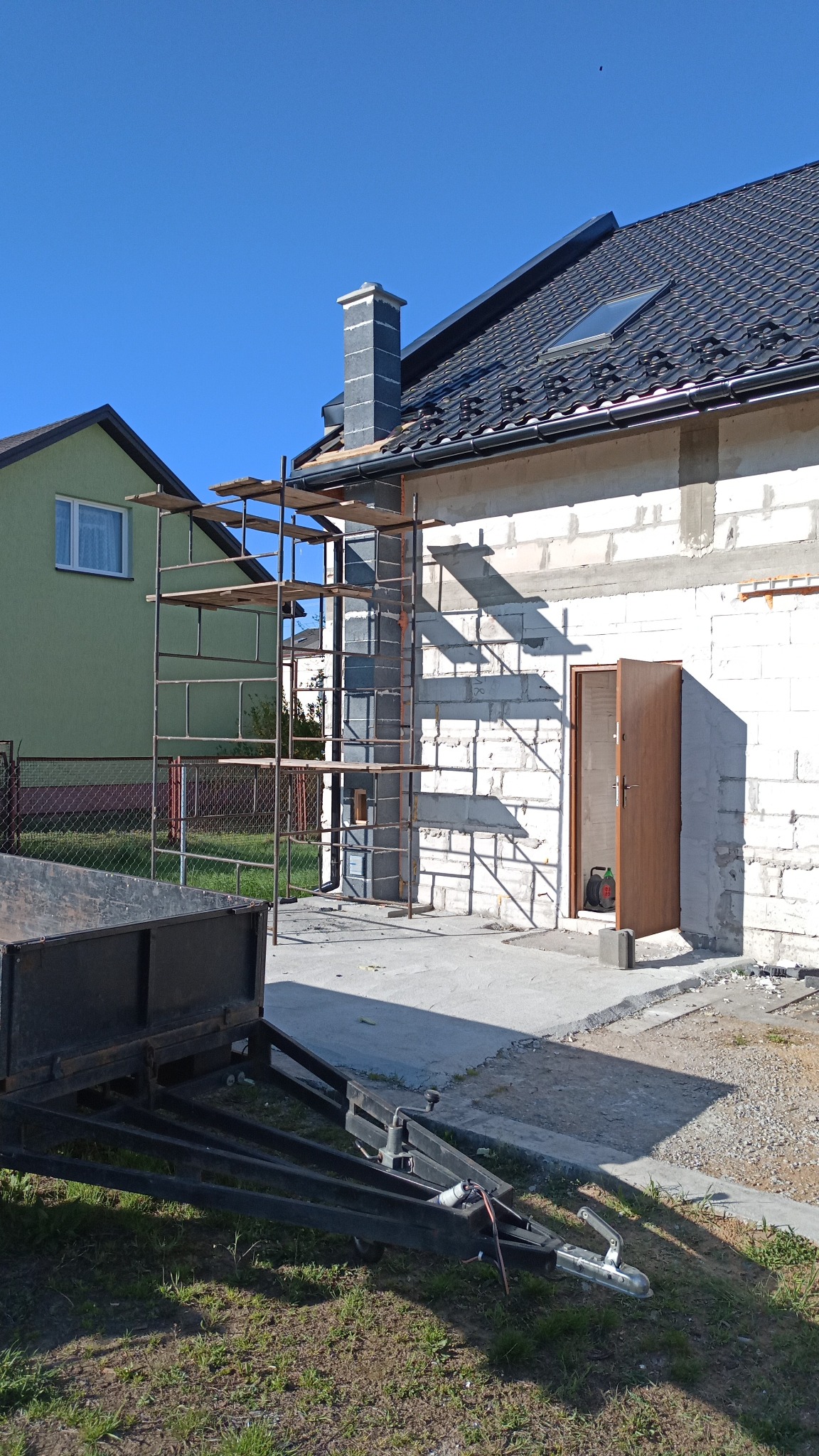 Budowa domu z białych bloczków, widoczne rusztowanie przy kominie, dach z czarnej dachówki. Przyczepka na trawie. Dom w trakcie budowy w słoneczny dzień.