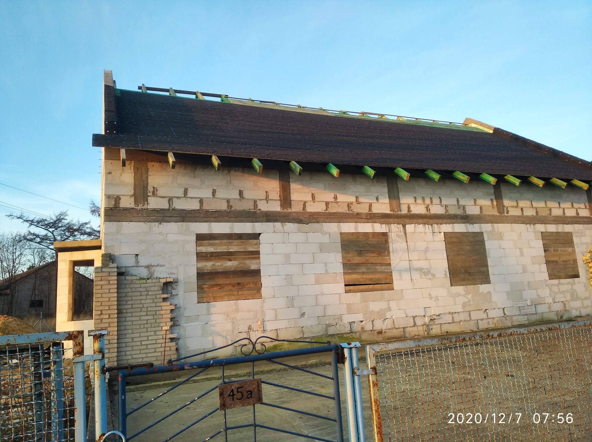 Niedokończony budynek z białych bloczków, z deskami zamiast okien, widoczne elementy dachu i brama z numerem 45a. W tle drzewa i niebo.