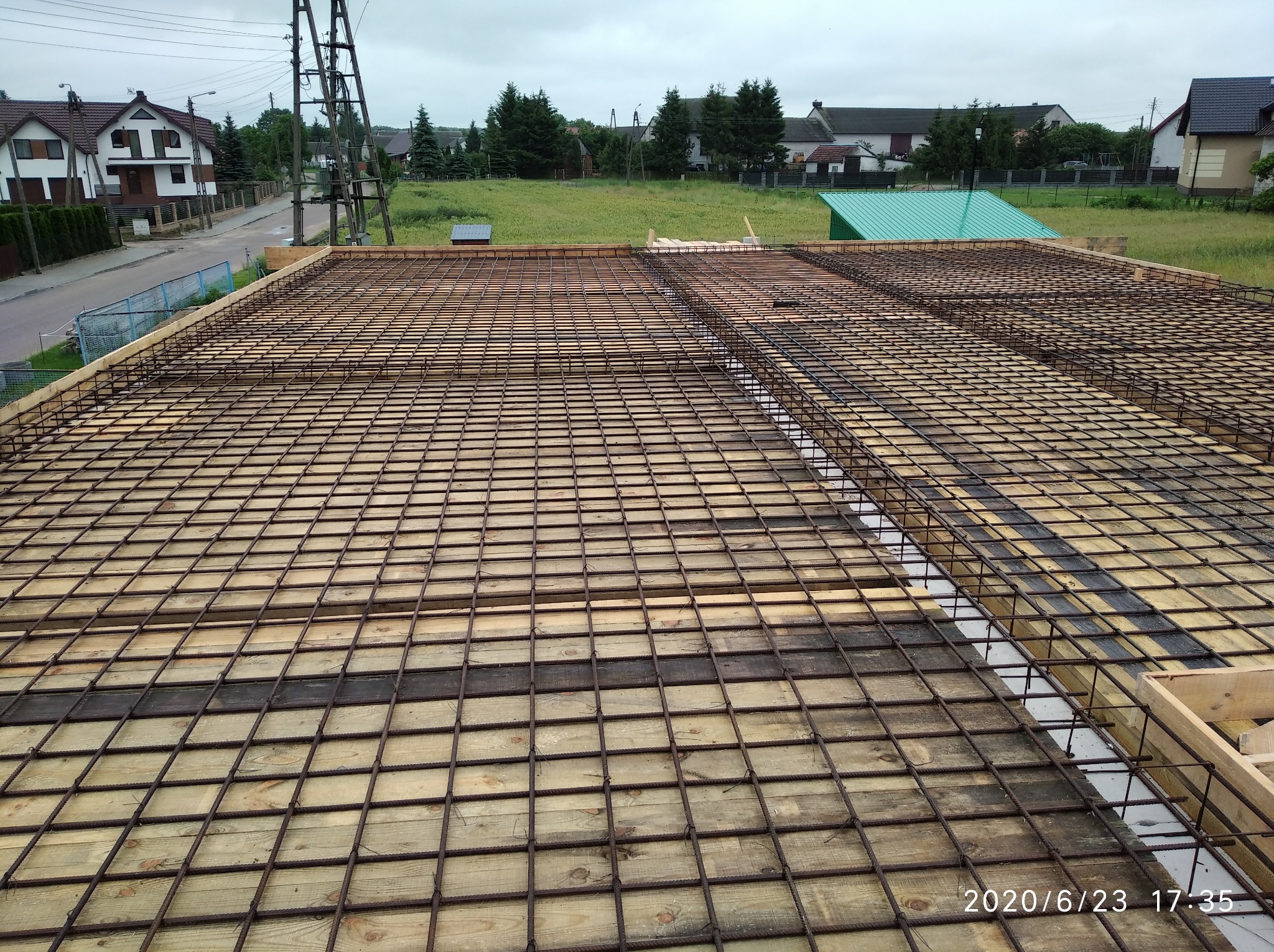 Zbrojenie stropu na deskowaniu z widocznymi prętami i drewnianymi elementami konstrukcyjnymi. W tle zabudowa jednorodzinna i zieleń. Data wykonania zdjęcia w prawym dolnym rogu.