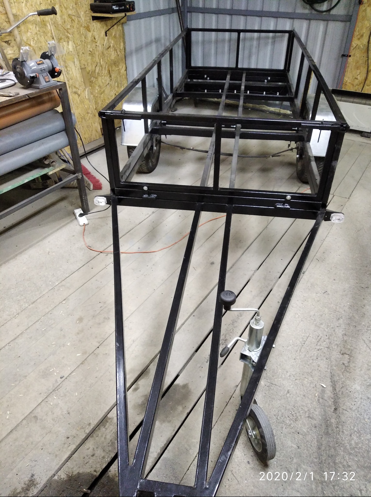 Metalowa konstrukcja przyczepy w warsztacie, widoczne spawane ramy, koła i elementy mocujące. Oświetlenie warsztatowe, narzędzia w tle.