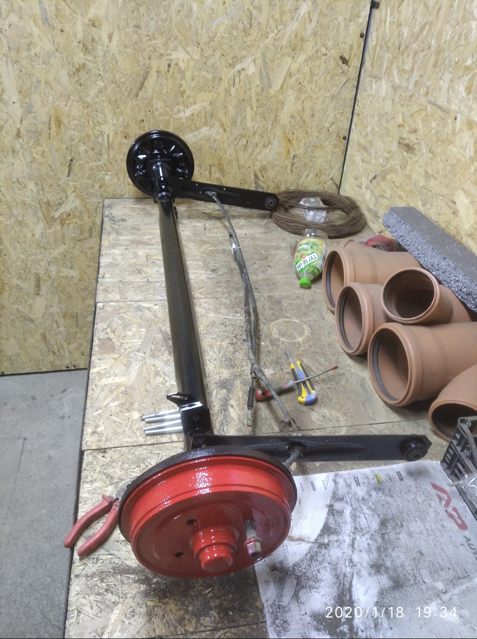 Odnowiona oś z czerwonym bębnem hamulcowym i czarnym ramieniem na warsztatowym stole, obok narzędzi i rur kanalizacyjnych. Widoczne ślady pracy.