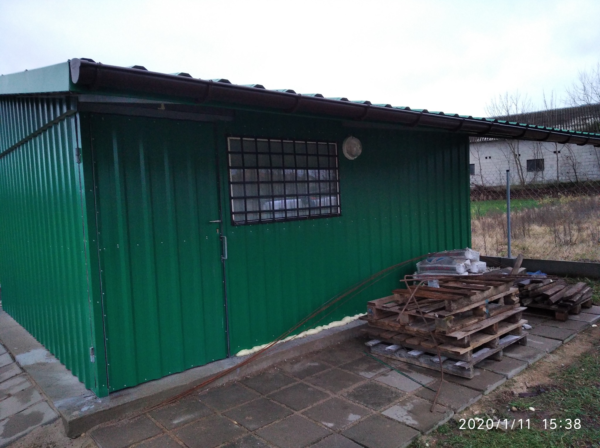 Zielony blaszak z oknem w kratkę i brązowymi rynnami, uszczelniony pianką montażową, z paletami obok, na tle szarego nieba i budynku w oddali.