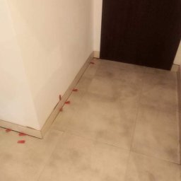 Tomasz Kochan - Ułożone płytki podłogowe w rogu pomieszczenia, z czerwonymi krzyżykami dystansowymi, widoczne przy listwie przypodłogowej i brązowych drzwiach.