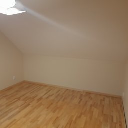Tomasz Kochan - Jasne panele podłogowe w pokoju na poddaszu, świeżo pomalowane ściany w odcieniach beżu, widoczna lampa sufitowa. Ukończone prace wykończeniowe.