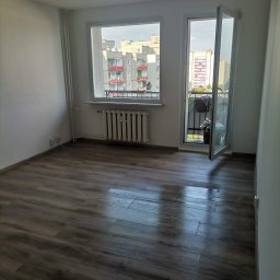 Tomasz Kochan - Pokój po remoncie: panele podłogowe w odcieniach brązu, białe ściany, okno z widokiem na budynki. Otwarty balkon z widocznymi przedmiotami.