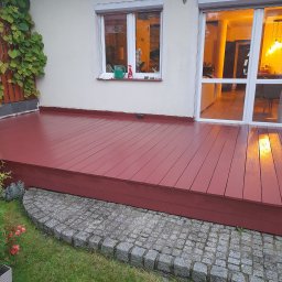Tomasz Kochan - Czerwony, świeżo pomalowany drewniany taras przylegający do domu z oknem i przeszklonymi drzwiami balkonowymi, otoczony zielenią i kostką brukową.