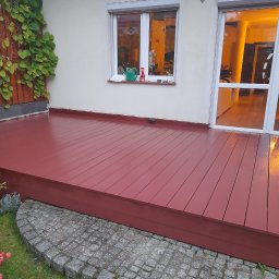 Tomasz Kochan - Czerwony, świeżo pomalowany drewniany taras przy domu z oknem i drzwiami balkonowymi. Widoczne rośliny pnące i kamienna obwódka. Ciepłe światło wewnątrz domu.