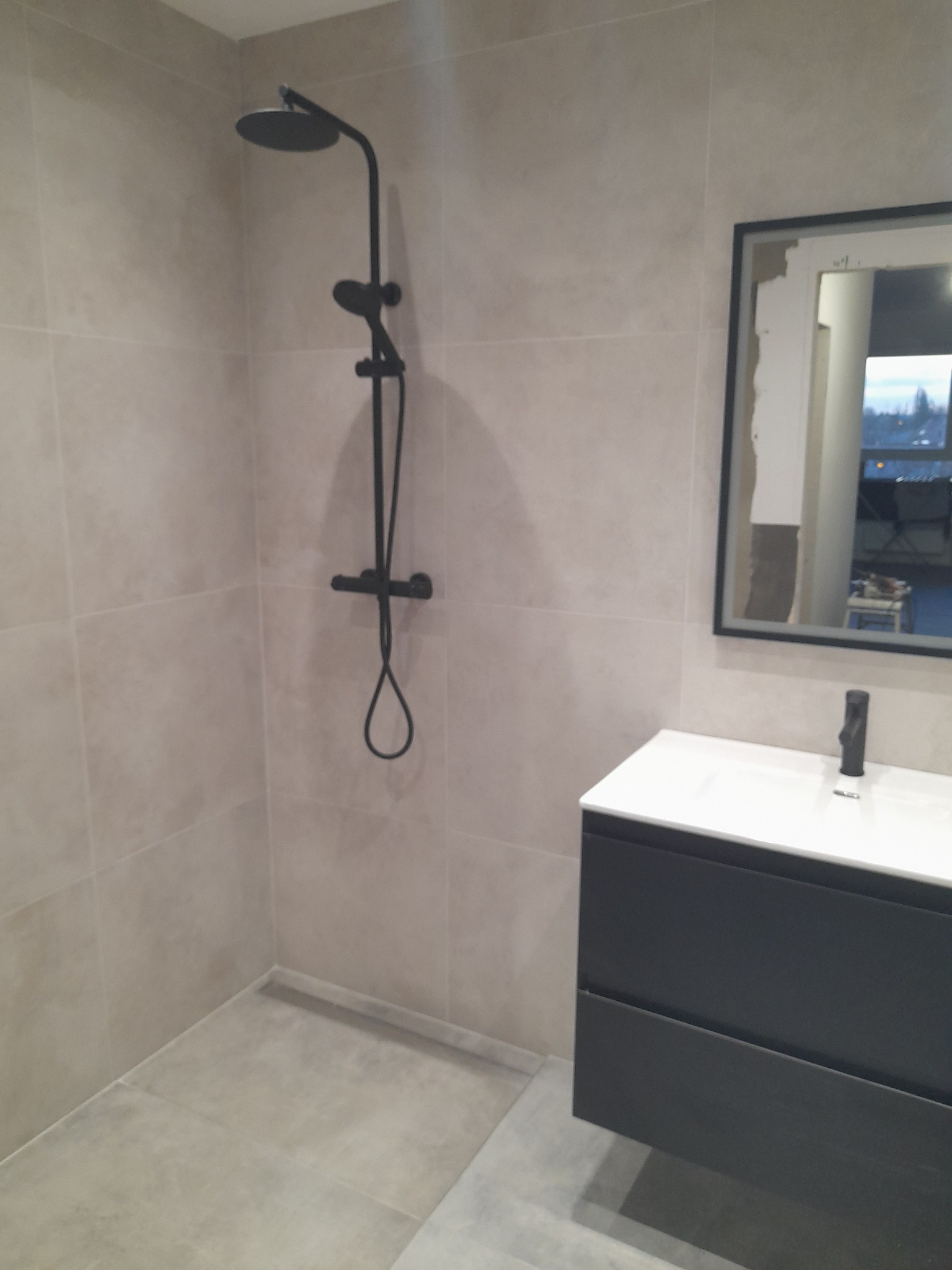 Wykończona łazienka z beżowymi płytkami, czarną armaturą i szafką pod umywalką. Widoczne lustro z czarną ramą i fragment widoku za oknem. Minimalistyczny design.