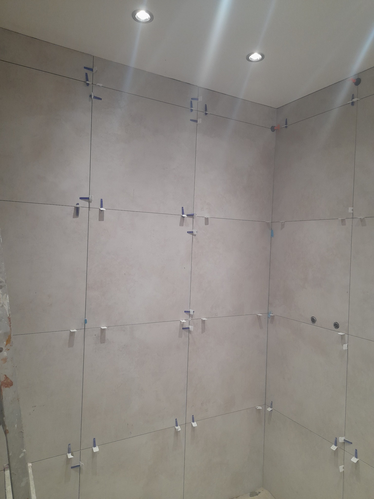 Ściana w trakcie układania płytek, z widocznymi krzyżykami dystansowymi i systemem poziomowania. Jasne płytki o delikatnej fakturze, oświetlenie sufitowe.