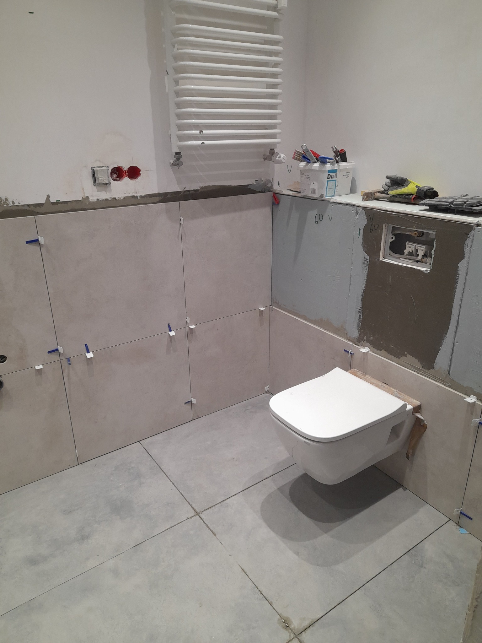 Wnętrze łazienki w trakcie remontu. Widoczne płytki na ścianach z krzyżykami dystansowymi, podwieszana toaleta i grzejnik drabinkowy. Częściowo położone płytki podłogowe.