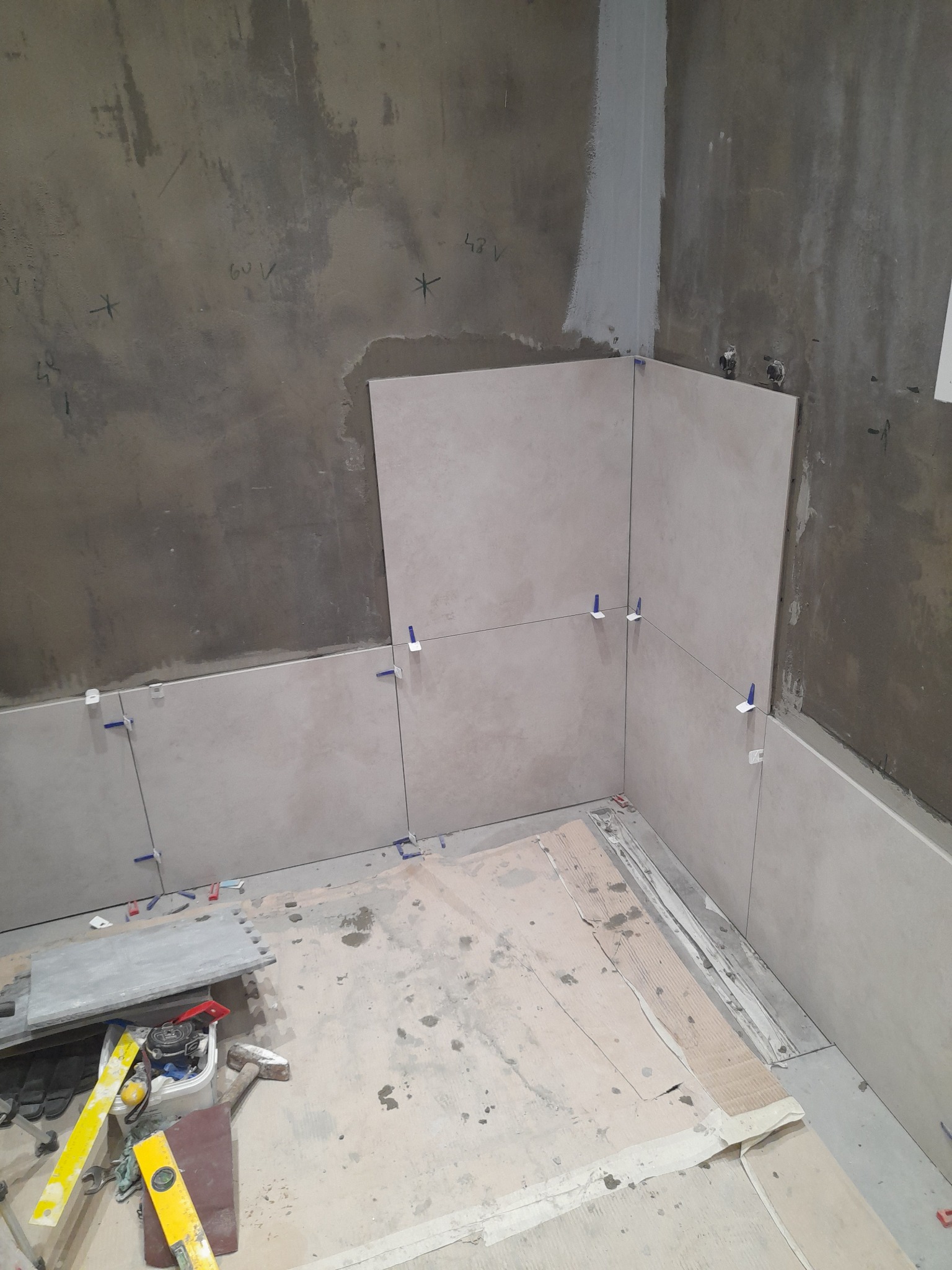 Układanie płytek w rogu pomieszczenia z użyciem krzyżyków dystansowych. Widoczne narzędzia i materiały. Prace glazurnicze w Polkowicach w toku.