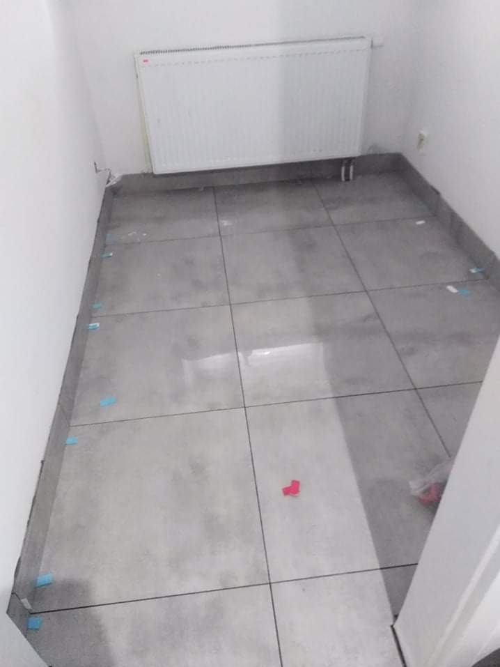 Szare płytki podłogowe w małym pomieszczeniu, z niebieskimi i czerwonymi elementami dystansowymi, grzejnik na ścianie. Widok z góry, ukończone układanie płytek.