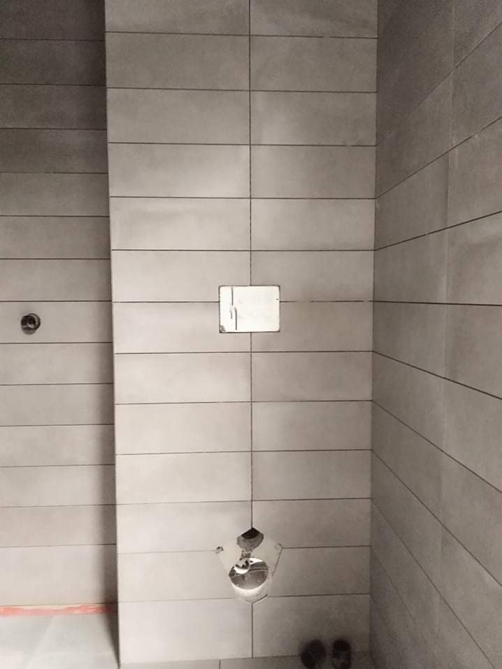 Ściana wyłożona szarymi, podłużnymi płytkami. Widoczne otwory na armaturę i instalację. Minimalistyczny design z wyraźnymi fugami. Ukończony etap prac wykończeniowych.