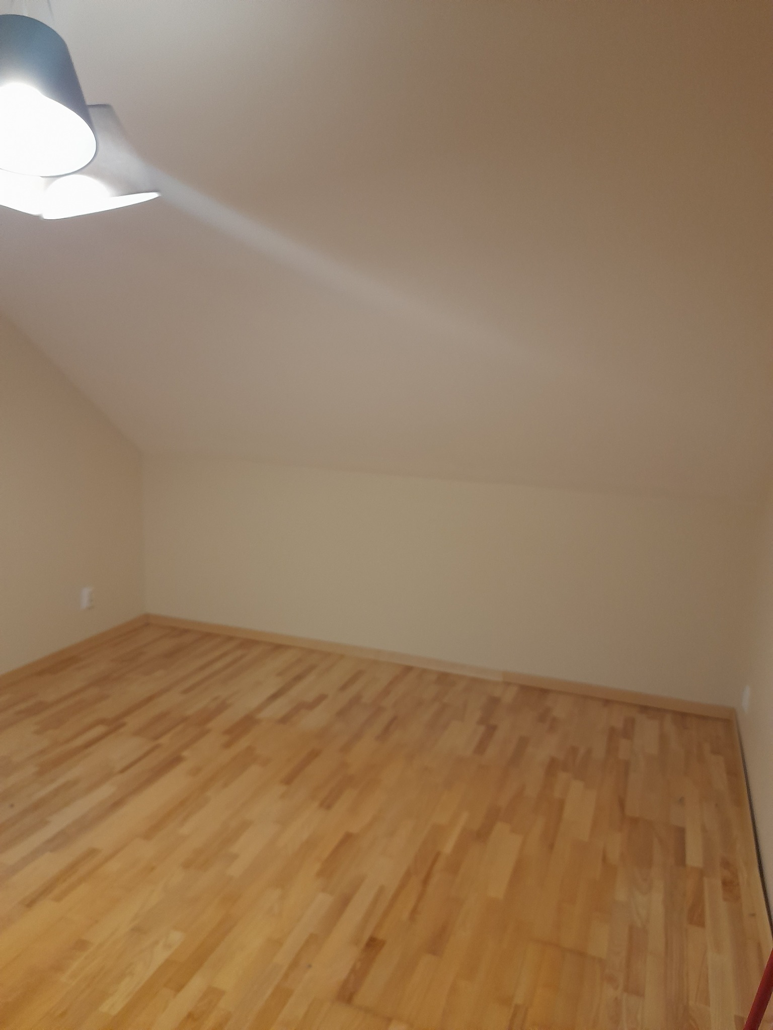 Jasne panele podłogowe w pokoju na poddaszu, świeżo pomalowane ściany w odcieniach beżu, widoczna lampa sufitowa. Ukończone prace wykończeniowe.