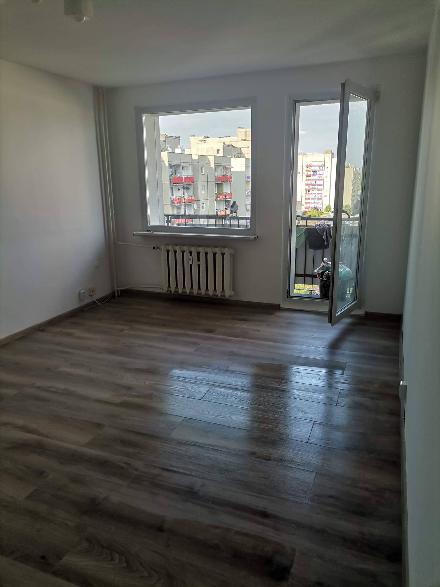 Pokój po remoncie: panele podłogowe w odcieniach brązu, białe ściany, okno z widokiem na budynki. Otwarty balkon z widocznymi przedmiotami.