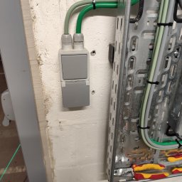 Elektro_Bud V.K. Warszawa - Szary, podwójny włącznik natynkowy z klapką, zamontowany na białej, chropowatej ścianie. Obok metalowa drabinka kablowa z przewodami w peszlach i narzędzia.