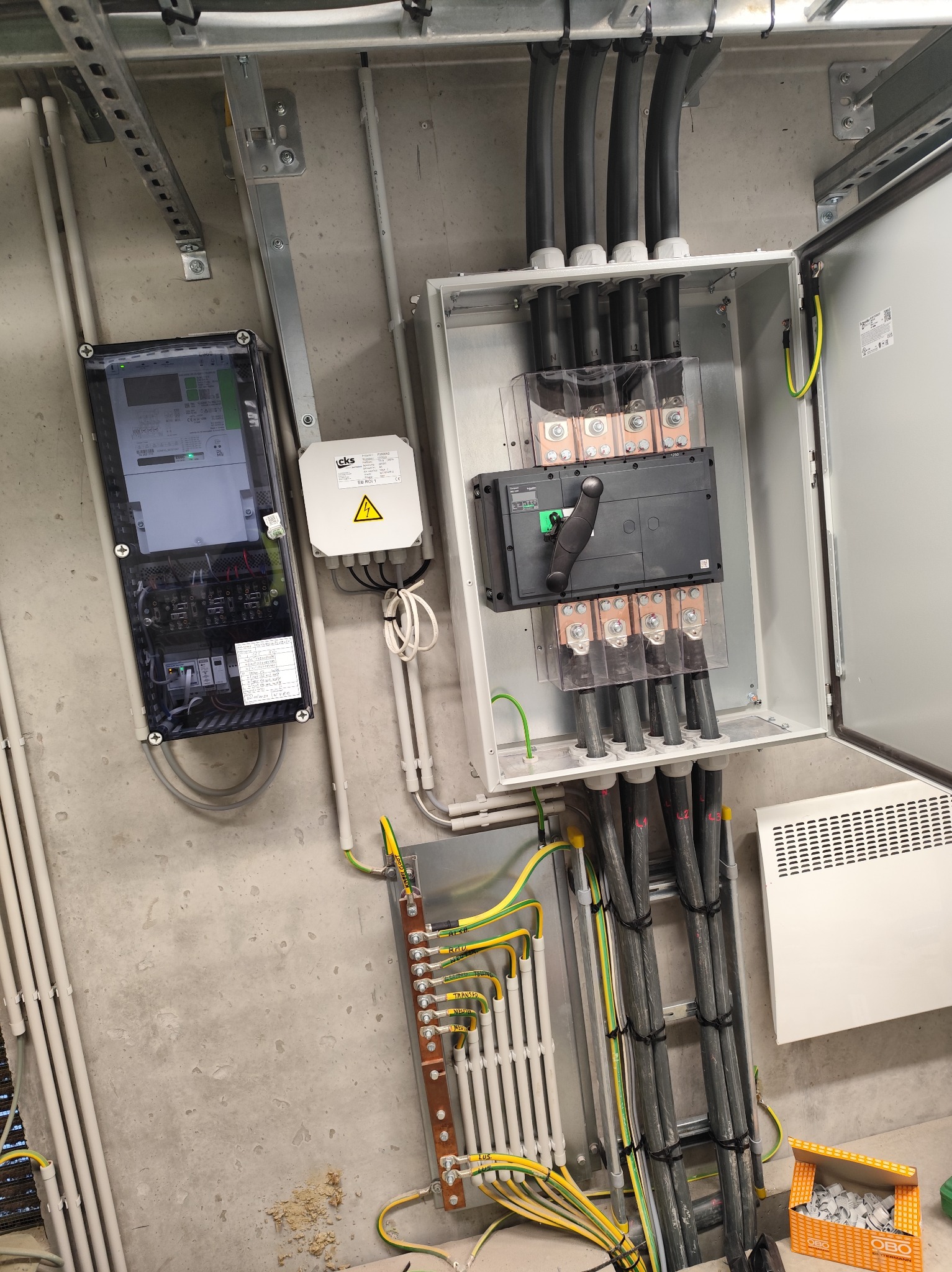Rozdzielnica elektryczna z podłączonymi kablami, widoczny licznik i zabezpieczenia. Instalacja w szarej, betonowej przestrzeni technicznej.