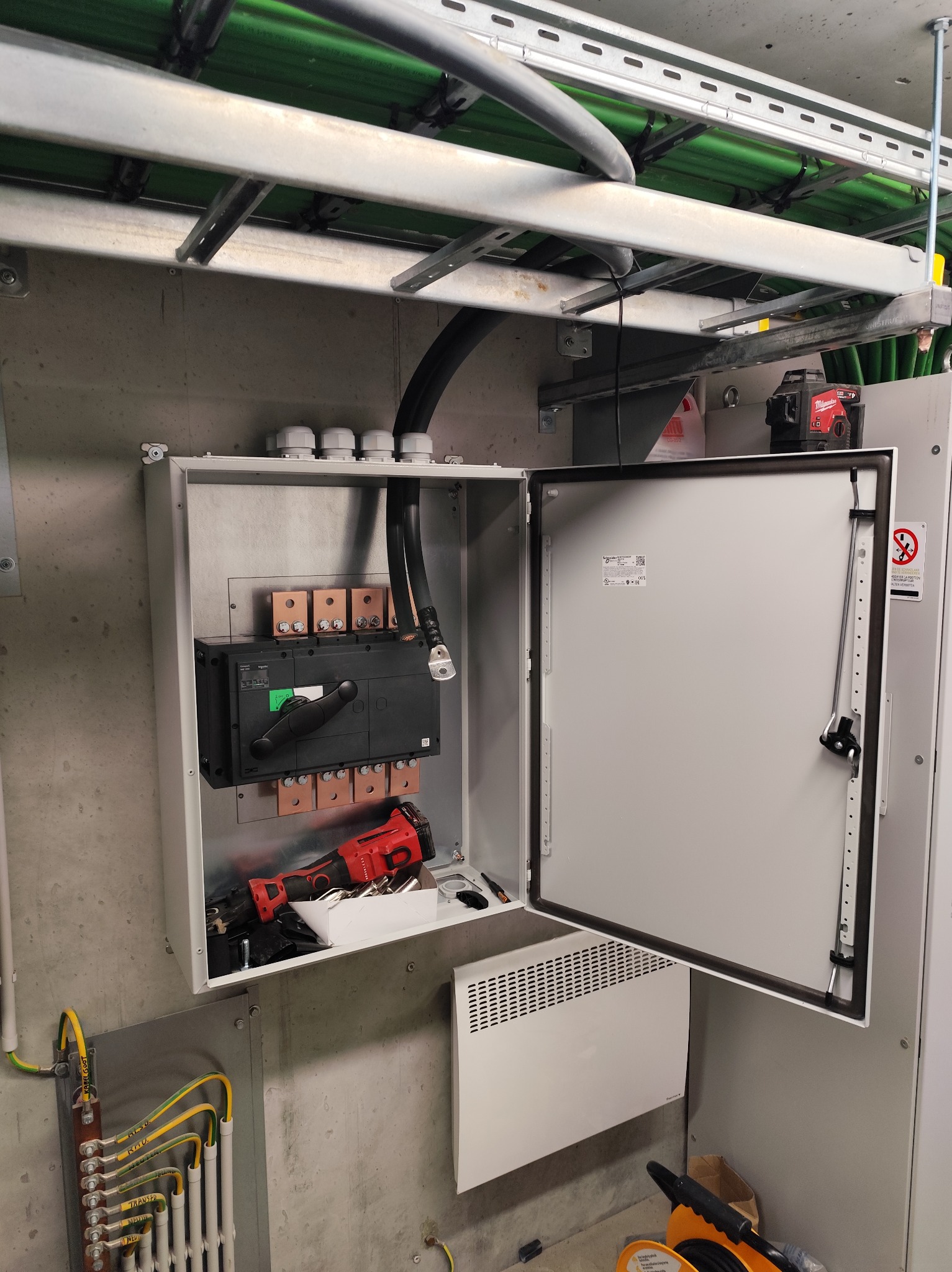 Otwarta szafa elektryczna z zamontowanym wyłącznikiem i narzędziami wewnątrz, widoczne kable i listwa zaciskowa. Szara ściana i elementy instalacji pod sufitem.