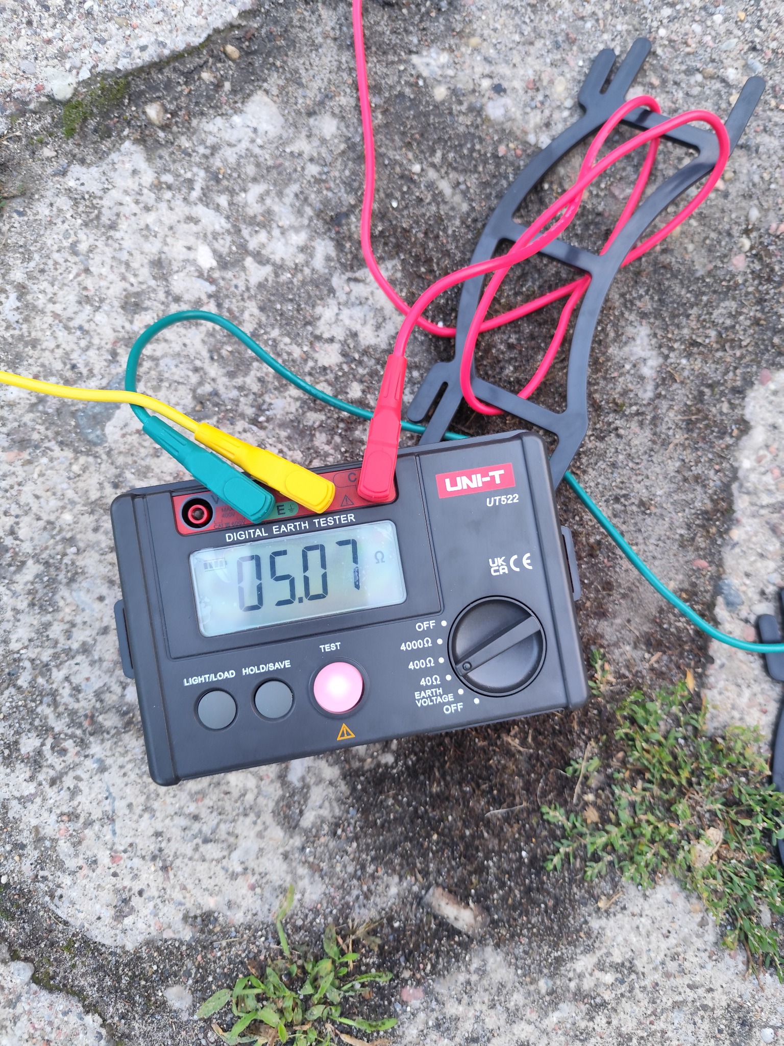 Miernik rezystancji uziemienia UNI-T UT522 z podłączonymi przewodami pomiarowymi na szorstkiej, betonowej powierzchni z zielenią. Wyświetlacz pokazuje wartość 05.07 Ω.