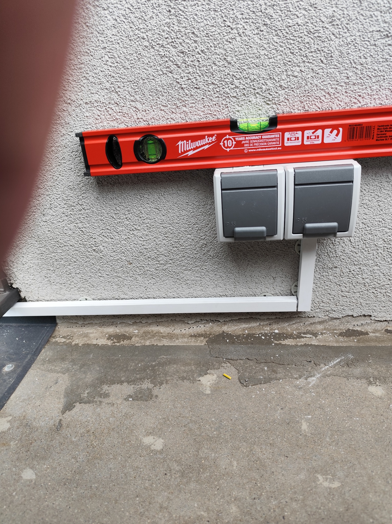 Dwa gniazda elektryczne IP55 zamontowane na szarej ścianie, poziomowane czerwoną poziomicą Milwaukee. Biała listwa przypodłogowa. Widoczny fragment podłoża.