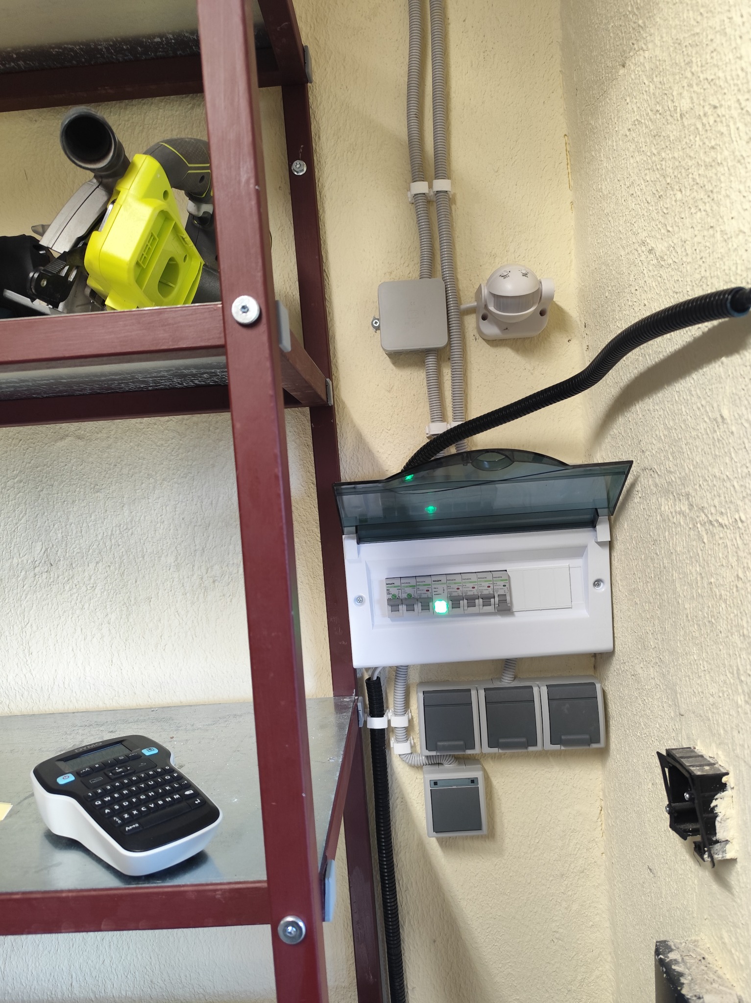 Rozdzielnia elektryczna z włączonymi bezpiecznikami, kable w peszlach, pistolet do etykiet Dymo na półce, narzędzia na regale. Żółta ściana z fakturą.