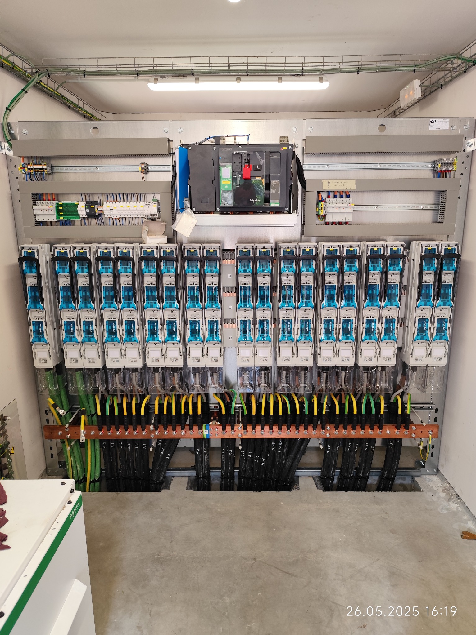 Rozbudowana szafa elektryczna z licznymi zabezpieczeniami, okablowaniem w kolorach żółtym i zielonym, zamontowana w pomieszczeniu o surowym wykończeniu.