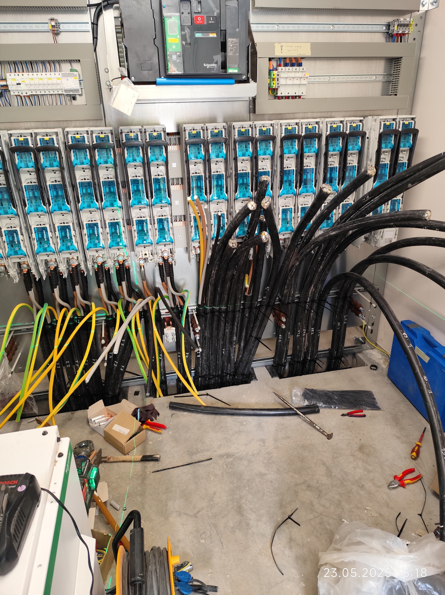 Rozdzielnia elektryczna w trakcie montażu: widoczne kable, listwy zaciskowe Wöhner i narzędzia. Oświetlenie górne, surowy beton na podłodze.