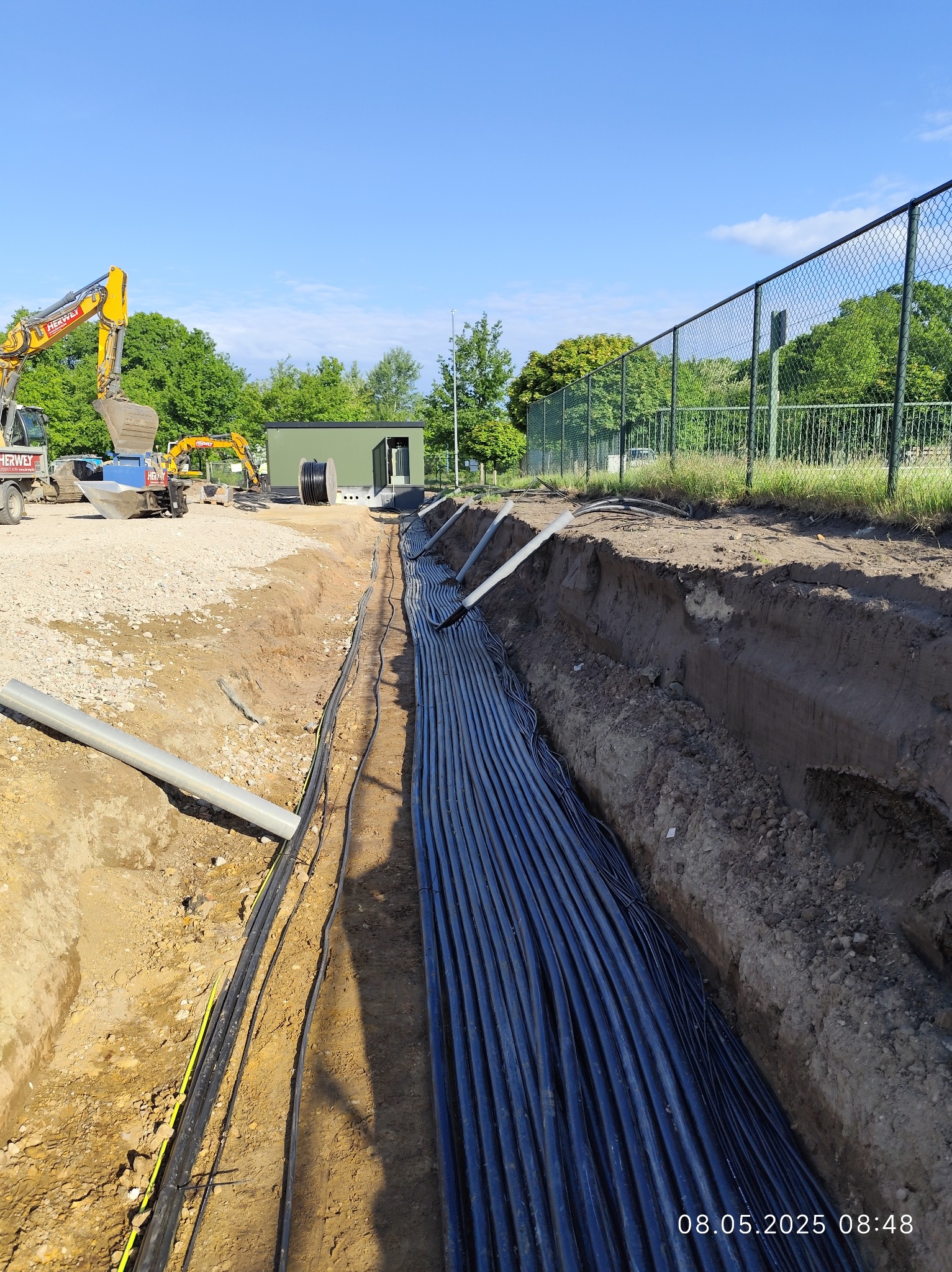 Wykop z ułożonymi kablami elektrycznymi w ziemi, widoczne rury osłonowe i maszyny budowlane w tle. Prace ziemne w słoneczny dzień.