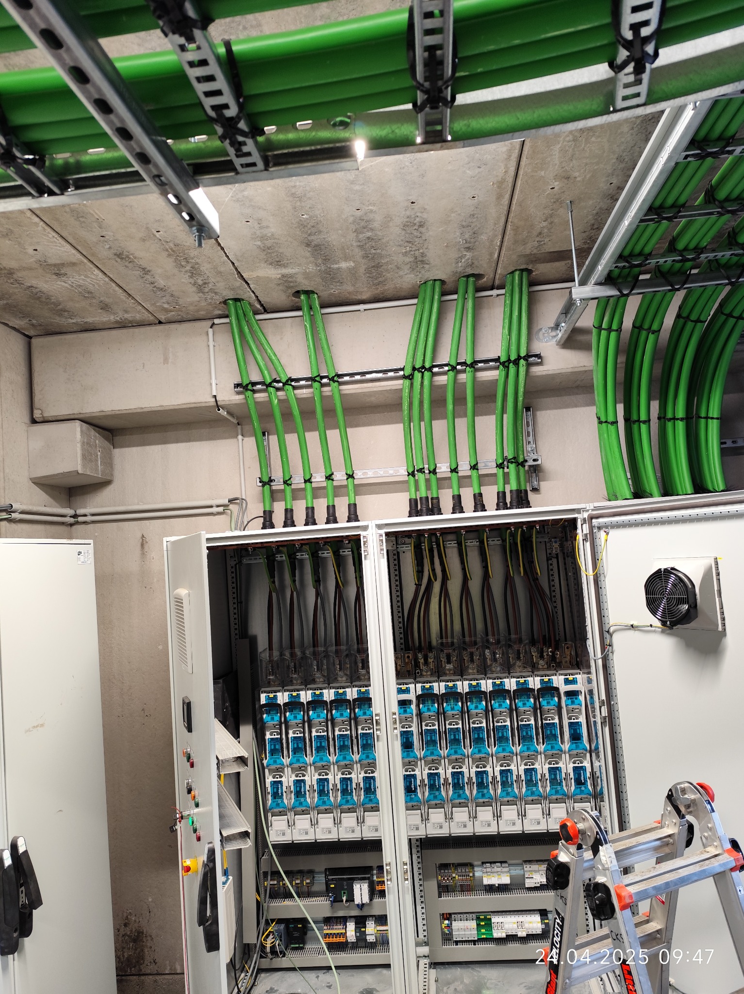 Rozdzielnia elektryczna z zielonymi rurami ochronnymi kabli, widoczne zabezpieczenia i aparatura. Drabina obok szafy. Instalacja w budynku przemysłowym.