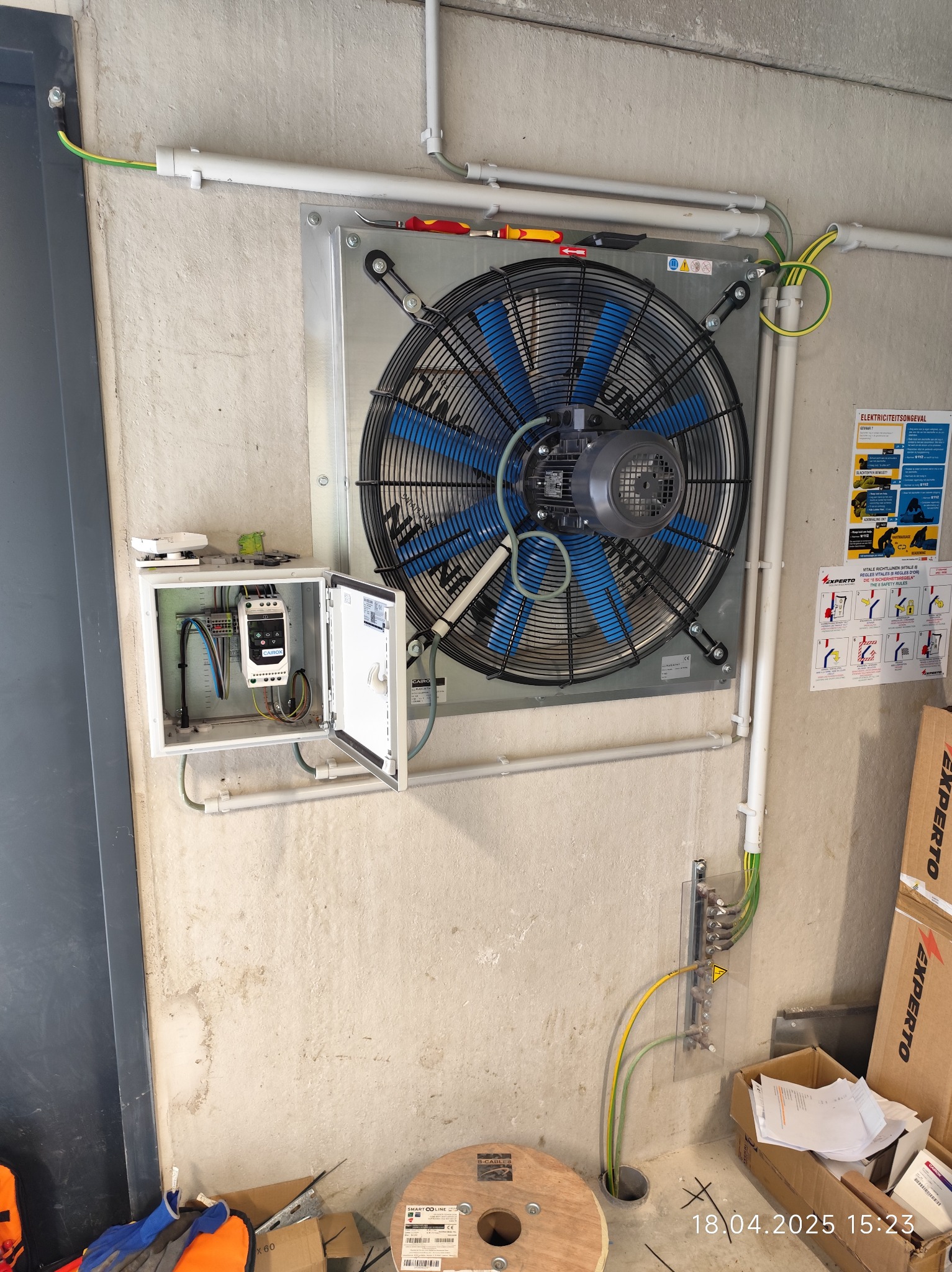 Wentylator przemysłowy z niebieskimi łopatkami, zamontowany na betonowej ścianie, z widoczną skrzynką elektryczną i okablowaniem. Widoczne elementy instalacji uziemiającej.