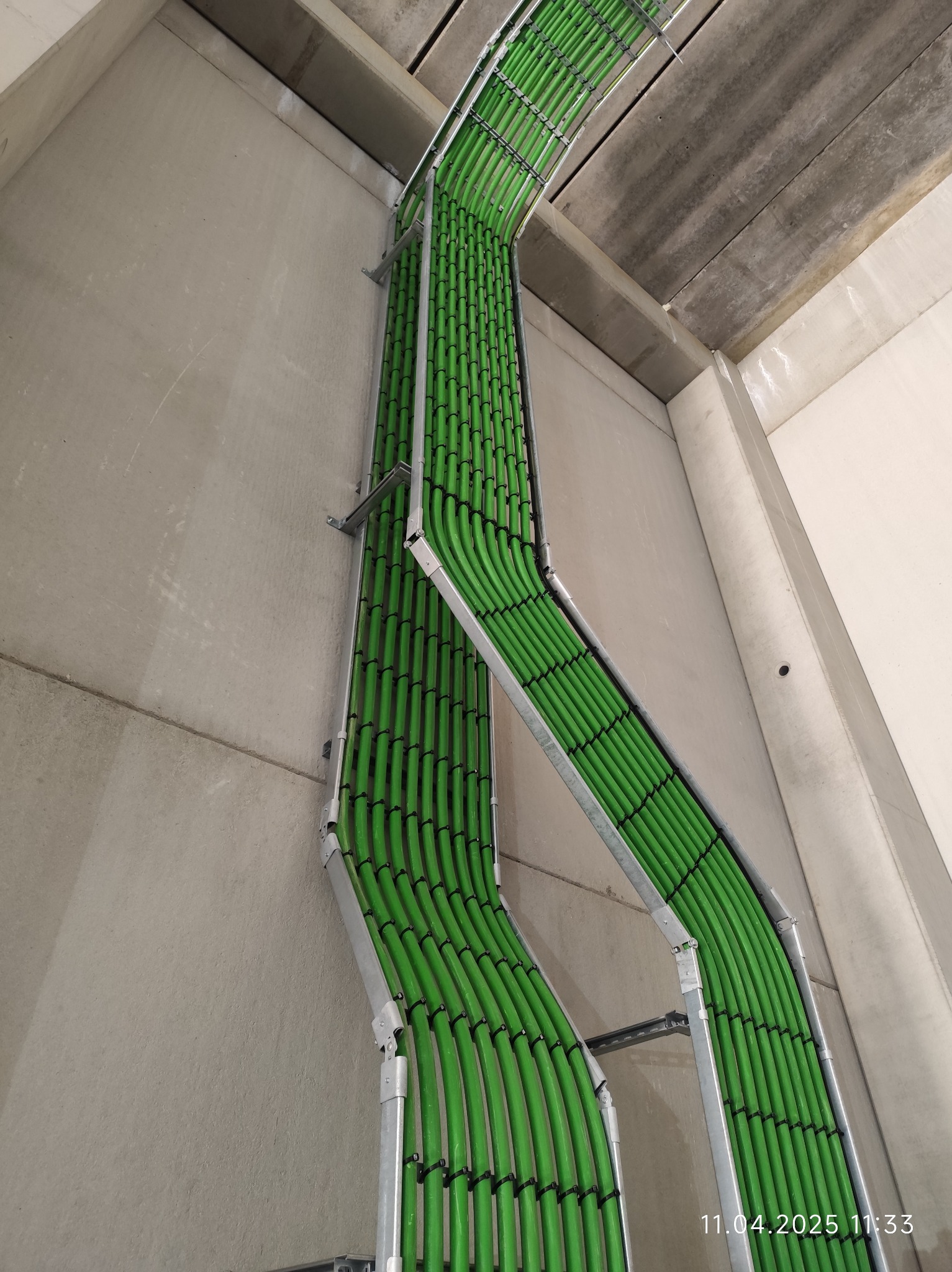 Równolegle ułożone, zielone kable elektryczne w metalowych korytach, zamocowane do surowej, betonowej ściany. Widok z dołu, ukazujący precyzję instalacji.