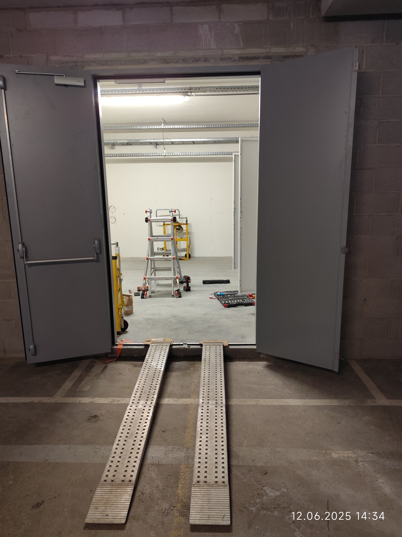 Otwarta, szara, metalowa brama garażowa z rampami wjazdowymi. W tle widoczna drabina, narzędzia i oświetlenie sufitowe. Ceglana ściana po bokach bramy.