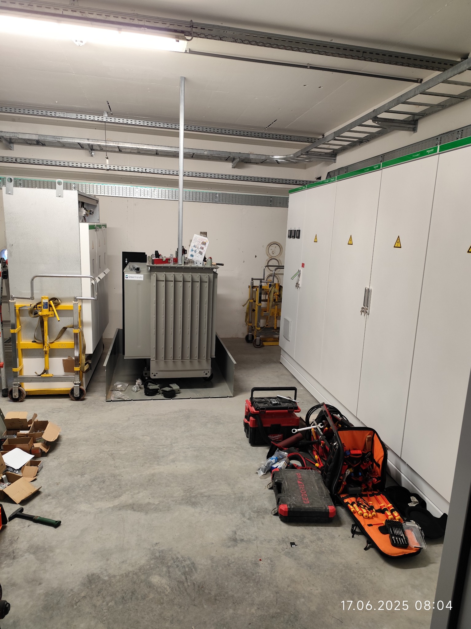 Wnętrze pomieszczenia z transformatorem, szafami elektrycznymi i narzędziami. Widoczne korytka kablowe pod sufitem. Na podłodze rozłożone skrzynki narzędziowe.