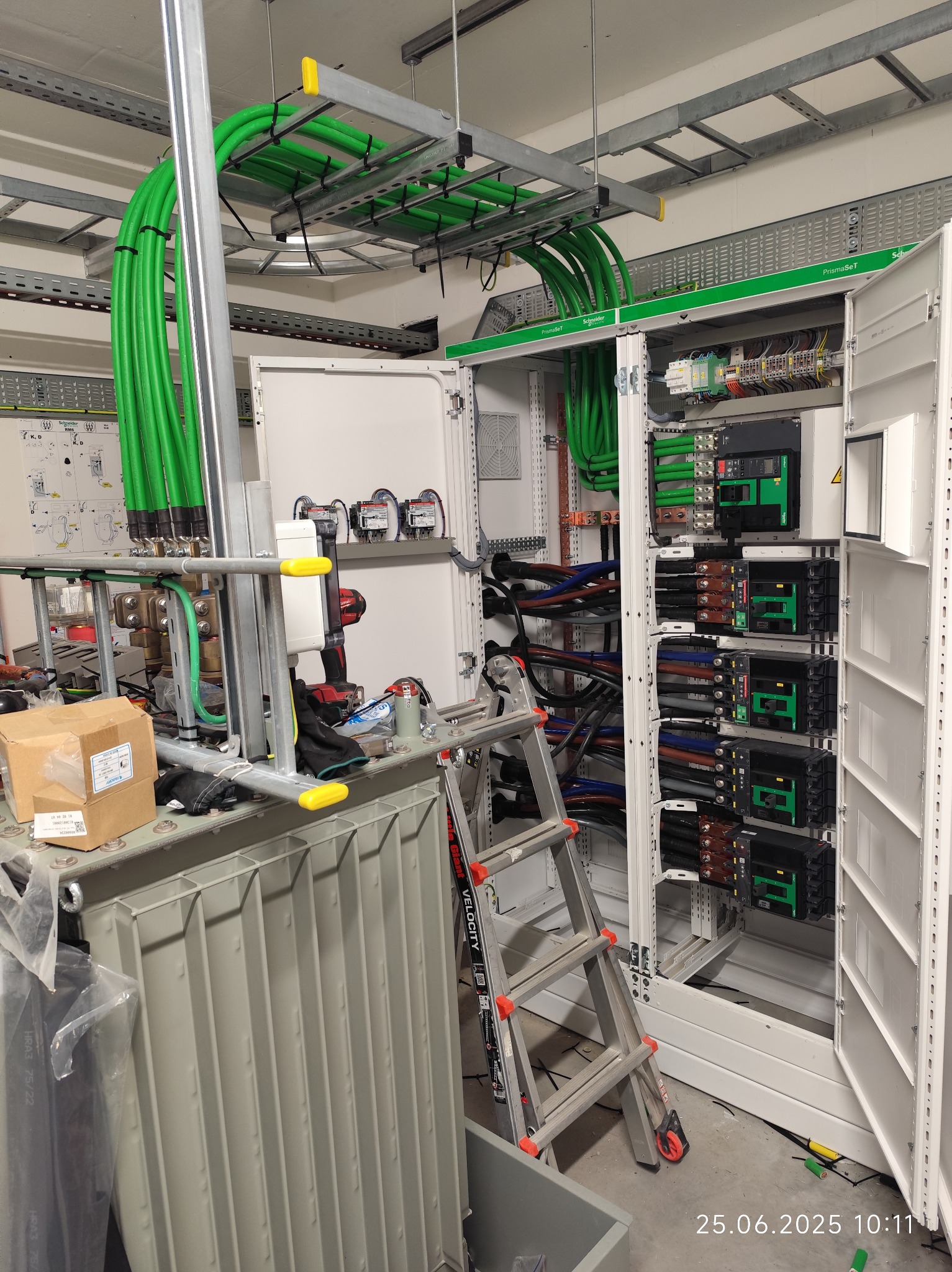 Otwarta szafa elektryczna PrismaSeT Schneider z zielonymi kablami, drabina i transformator w tle. Widoczne zabezpieczenia i okablowanie wewnątrz szafy.