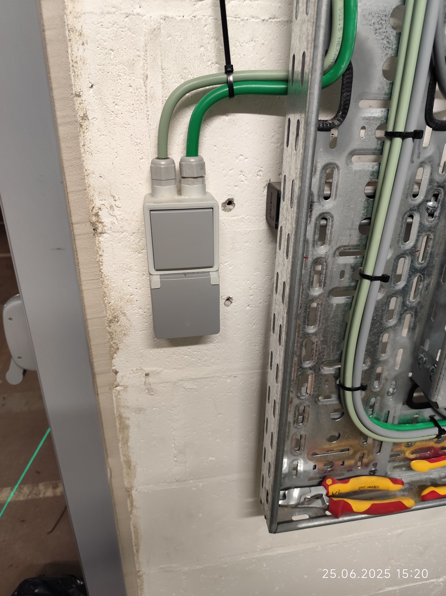 Szary, podwójny włącznik natynkowy z klapką, zamontowany na białej, chropowatej ścianie. Obok metalowa drabinka kablowa z przewodami w peszlach i narzędzia.