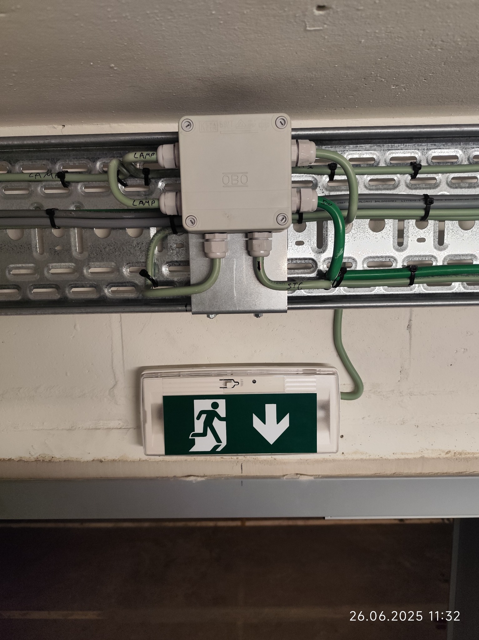 Szara puszka elektryczna OBO z przewodami na metalowej szynie, zamocowana nad znakiem ewakuacyjnym na białej ścianie. Widoczne kable oznaczone 'LAMP'.