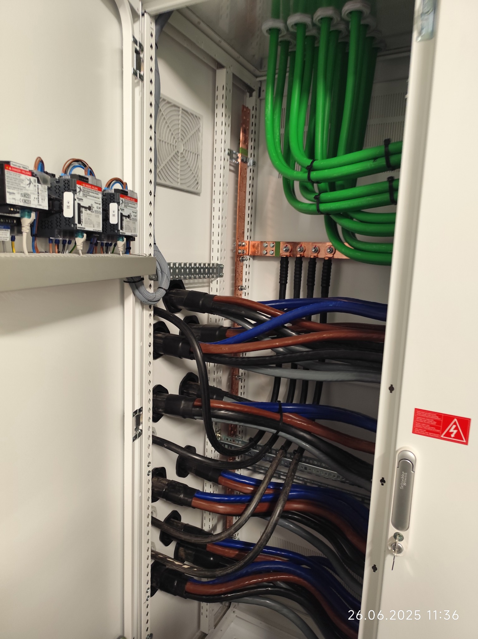 Wnętrze szafy elektrycznej z uporządkowanymi kablami w różnych kolorach (zielone, niebieskie, brązowe) i zabezpieczeniami. Widoczny wentylator i listwa zaciskowa.