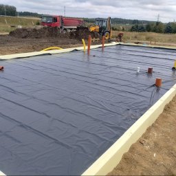 Piotr Bzówka - Przygotowany teren pod fundamenty z izolacją termiczną i folią hydroizolacyjną. W tle ciężarówka i koparka, widoczny pracownik.