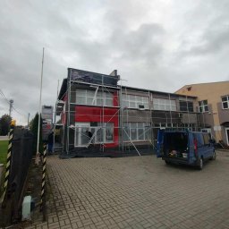 Piotr Bzówka - Budynek komercyjny w trakcie renowacji elewacji, otoczony rusztowaniami. Widoczny samochód dostawczy i fragment chodnika z kostki brukowej. Pochmurne niebo.