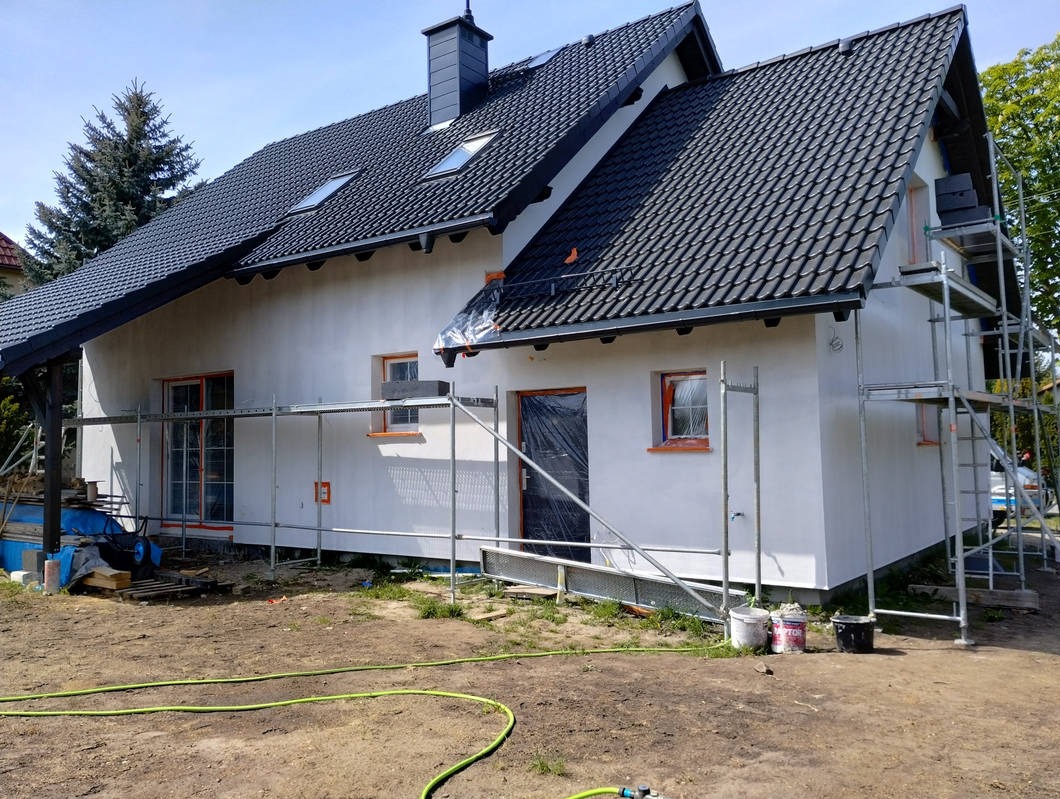 Dom z czarną dachówką w trakcie ocieplania elewacji, widoczne rusztowania, okna z pomarańczowymi ramami i częściowo zakryte folią. Na pierwszym planie wąż ogrodowy.