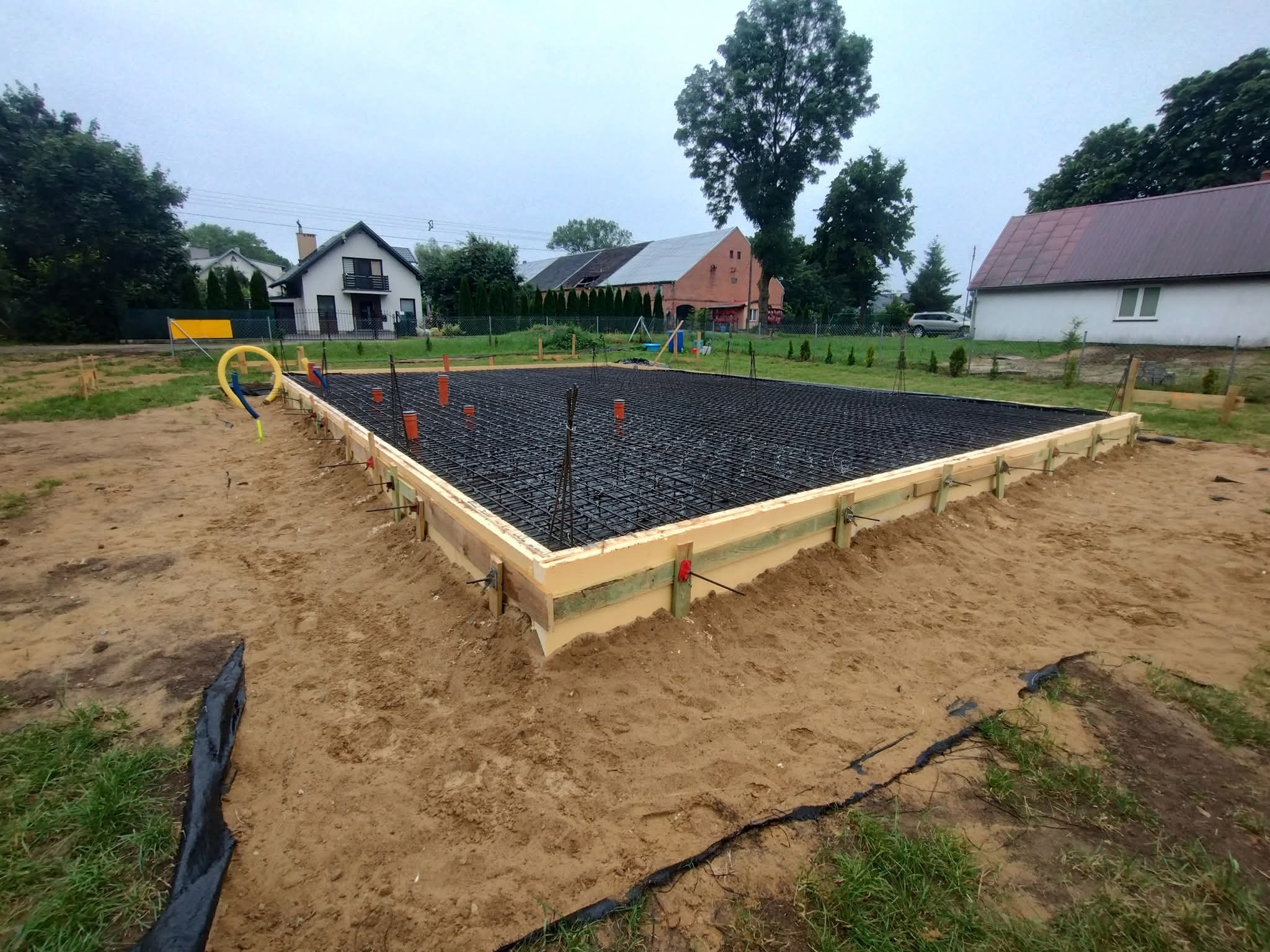 Zbrojenie fundamentów domu jednorodzinnego, widok z góry na przygotowany szalunek z izolacją i siatką zbrojeniową. W tle zabudowa jednorodzinna i zieleń.