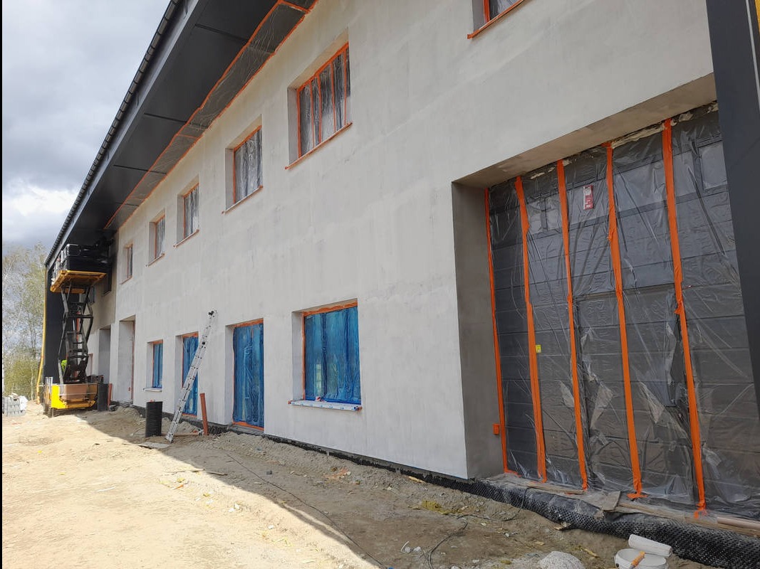Świeżo otynkowana elewacja budynku z zabezpieczonymi oknami i bramą folią malarską. Widoczna drabina i podnośnik koszowy. Prace wykończeniowe w toku.